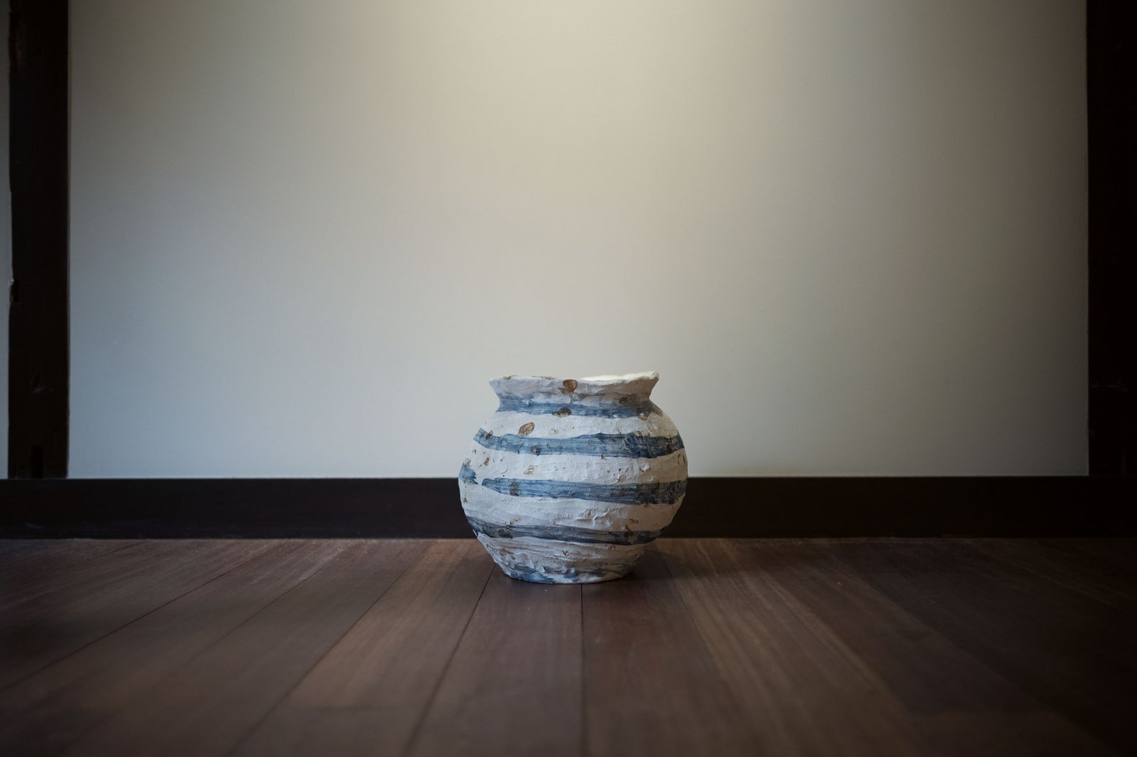 A white jar with blue bands on a dark wooden floor against a pale wall.