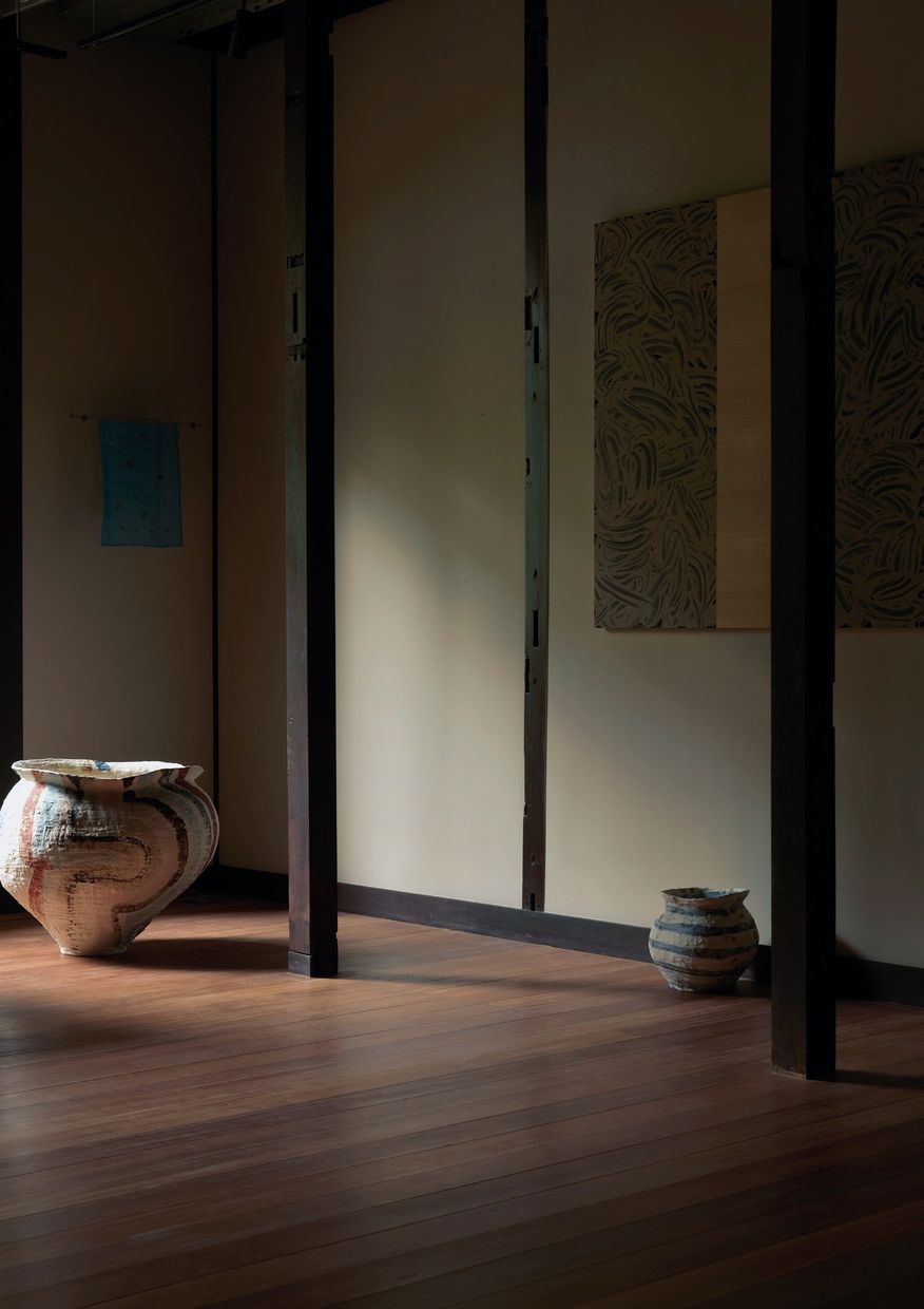 A dim room with a large striped jar in the corner, a small jar against the wall and a blue textile and a mud work hanging beyond.