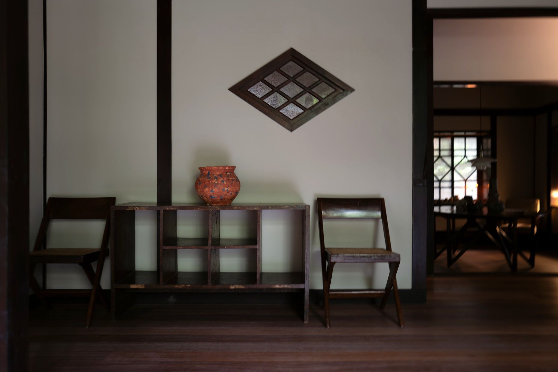 An orange jar on a low shelf between two wooden chairs, under a diamond-shaped lattice window.