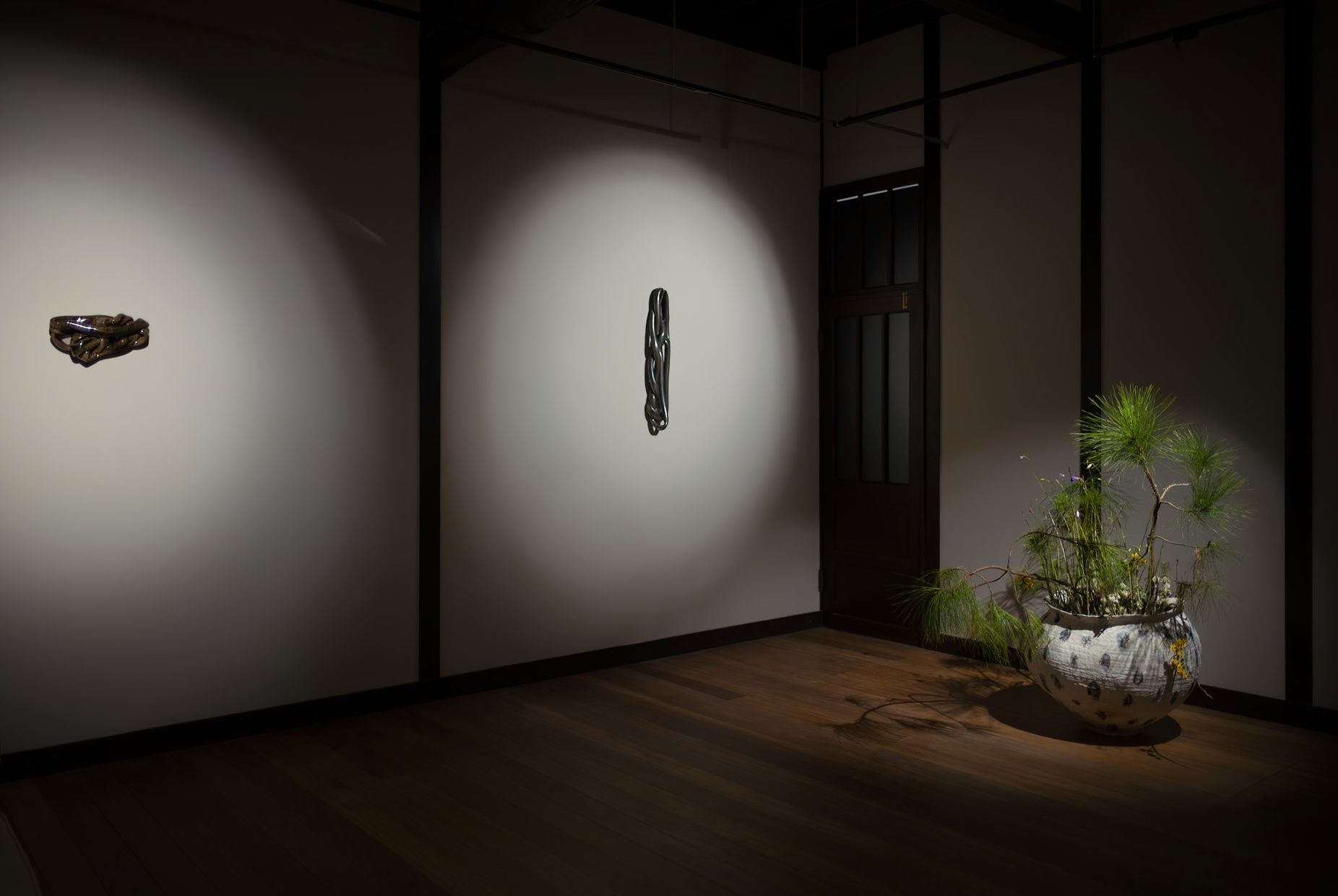 Two ceramic knots by Nicole Ondre lit on a pale wall, with a pine arrangement in a jar in the corner.