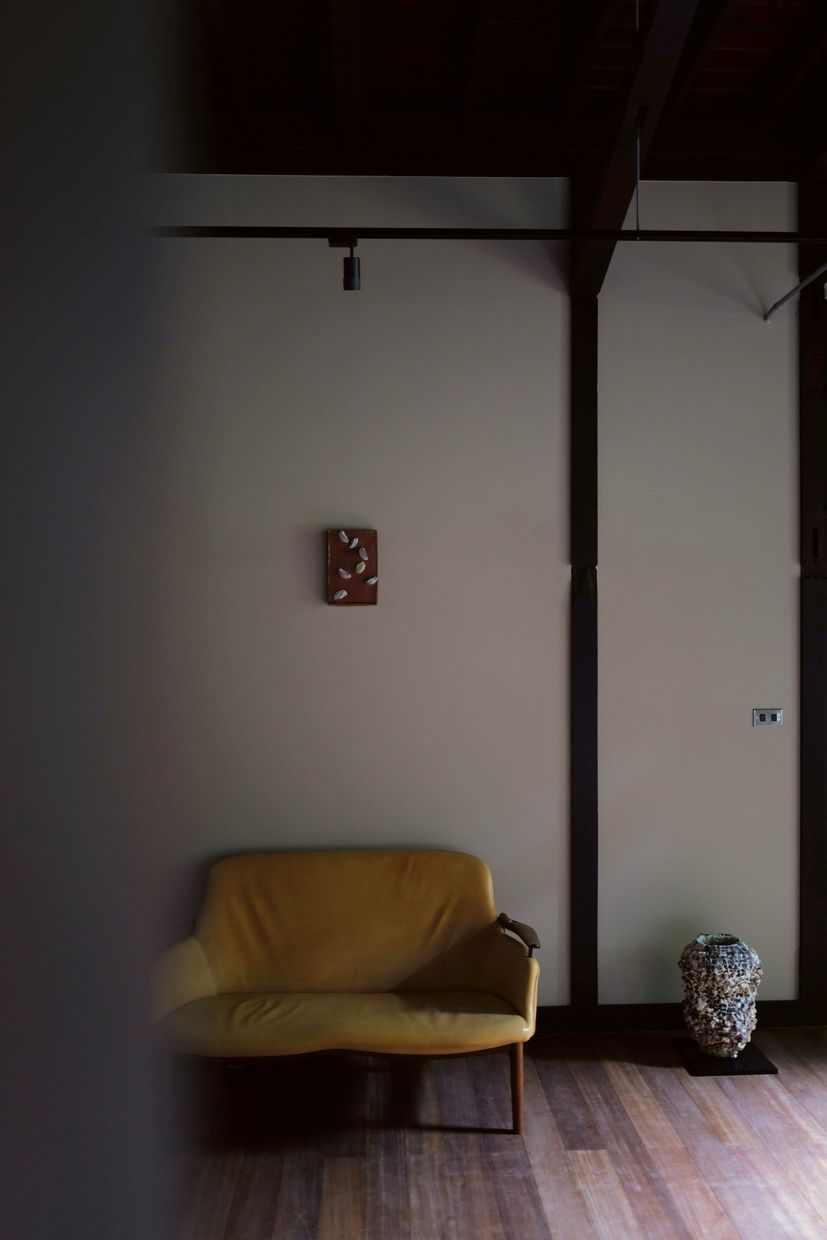 A yellow sofa below a small ceramic panel on a pale wall, with a mosaic vessel on a plinth beside a wooden pillar.