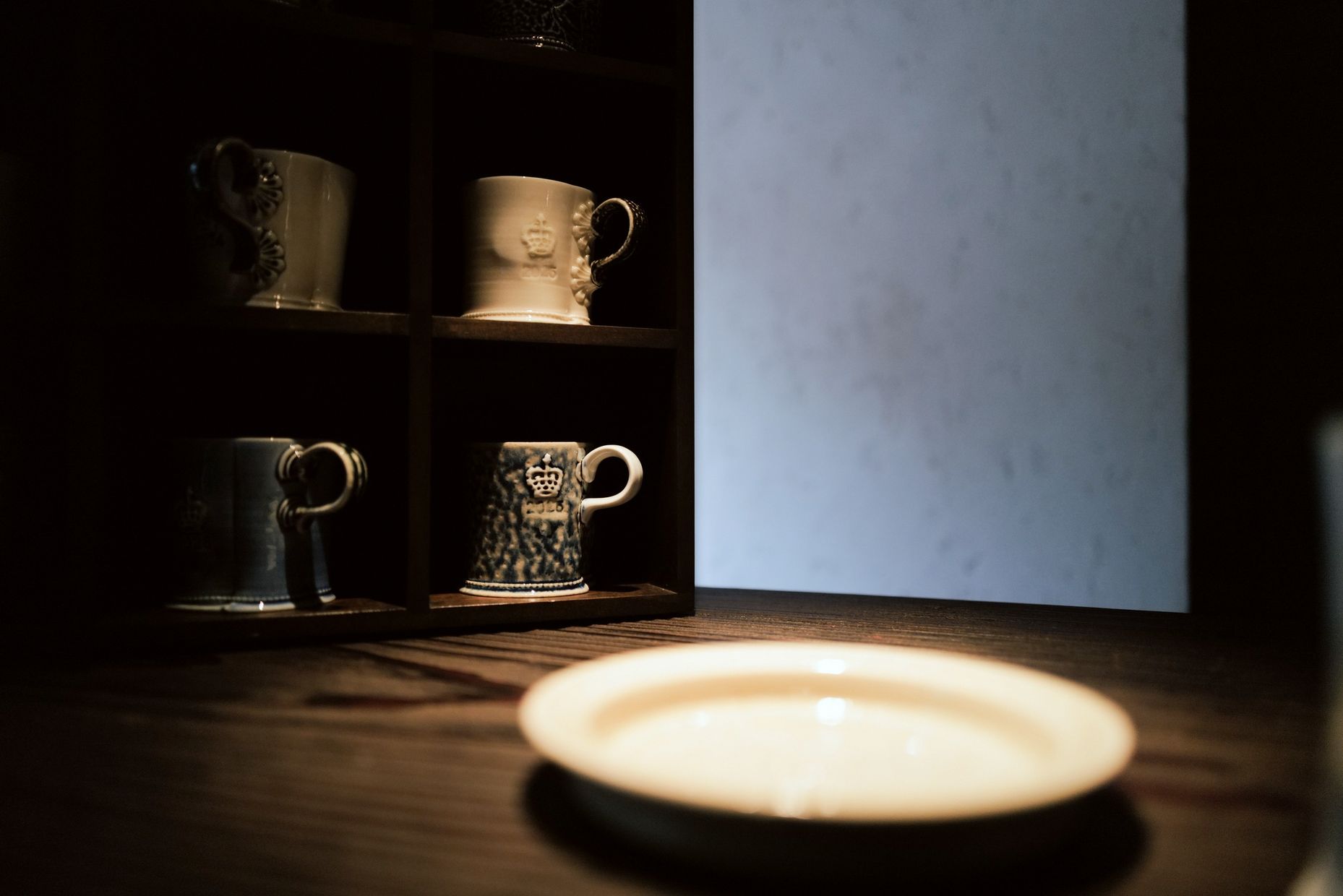 Salt-glazed mugs on dark wooden shelves, with a pale plate on the table in front.