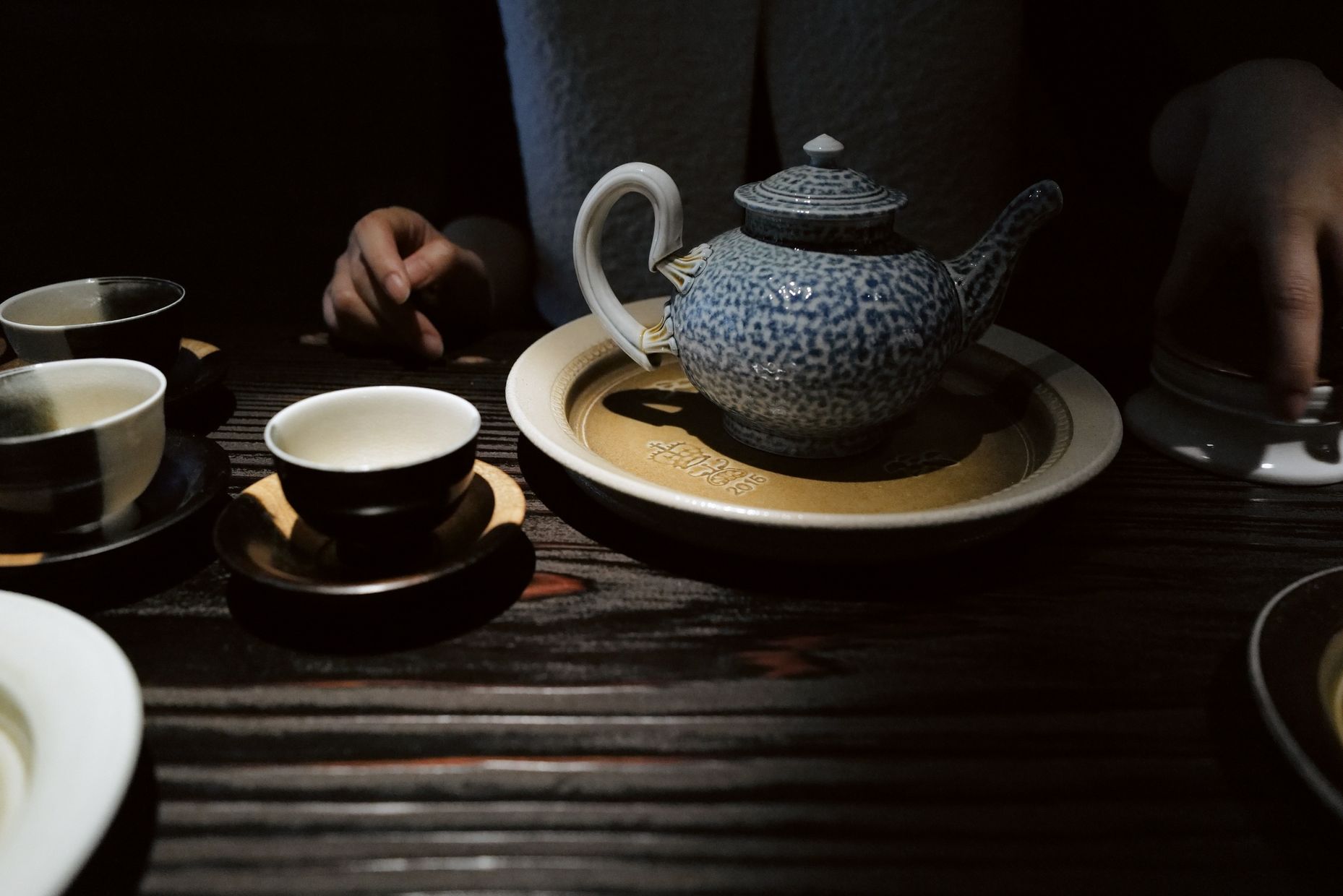 Guests at a dark wooden tea table set with a blue salt-glazed teapot on a plate, small cups and saucers.