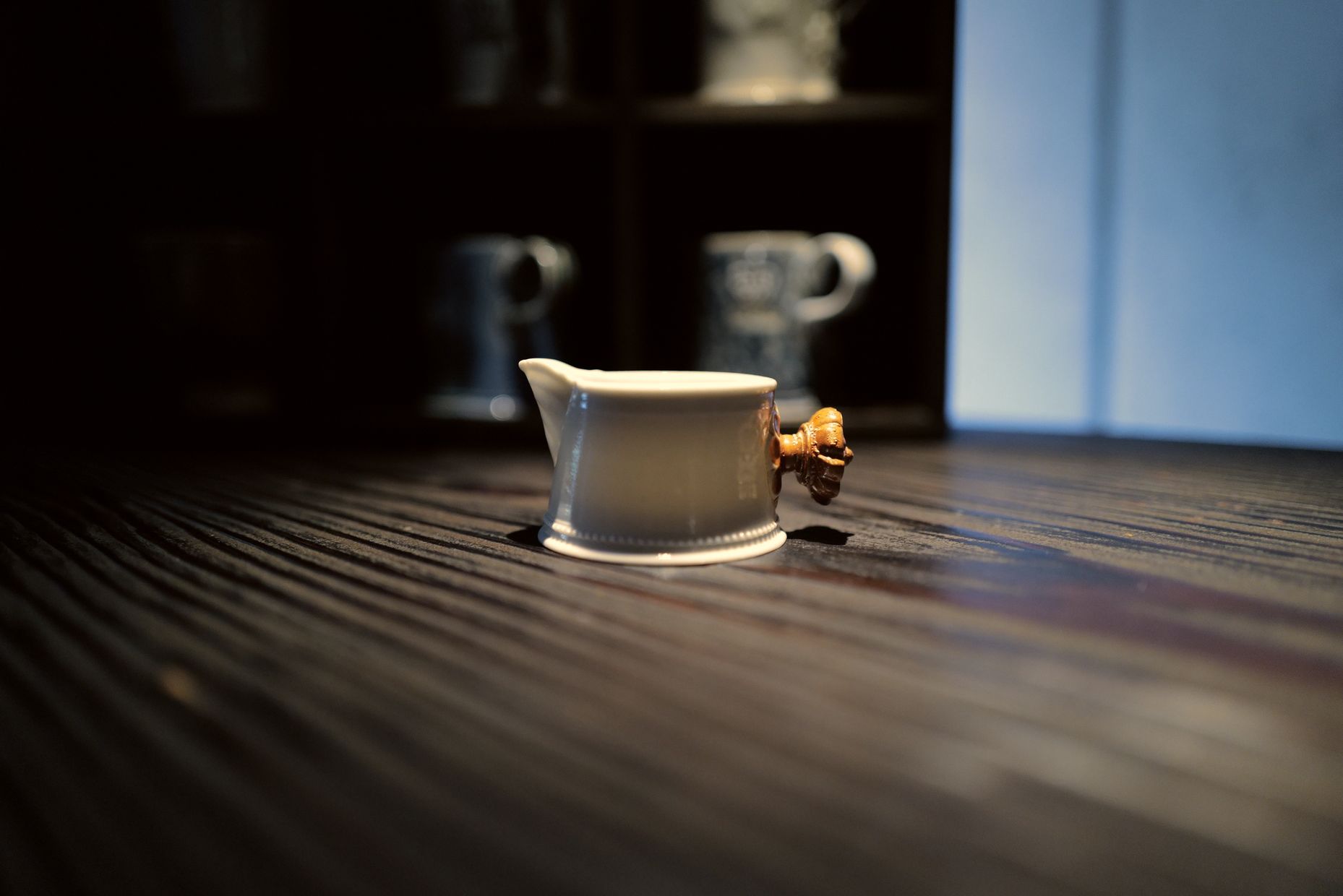 A white pouring jug with a carved wooden handle on a dark grooved table, mugs on shelves behind.