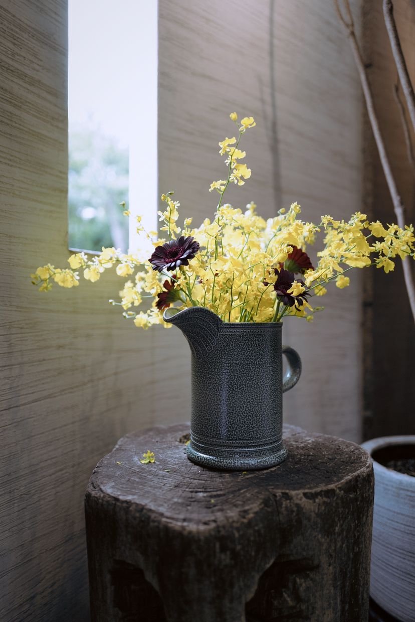 Yellow oncidium orchids and dark flowers in a blue-grey salt-glazed jug on a tree stump by a window.