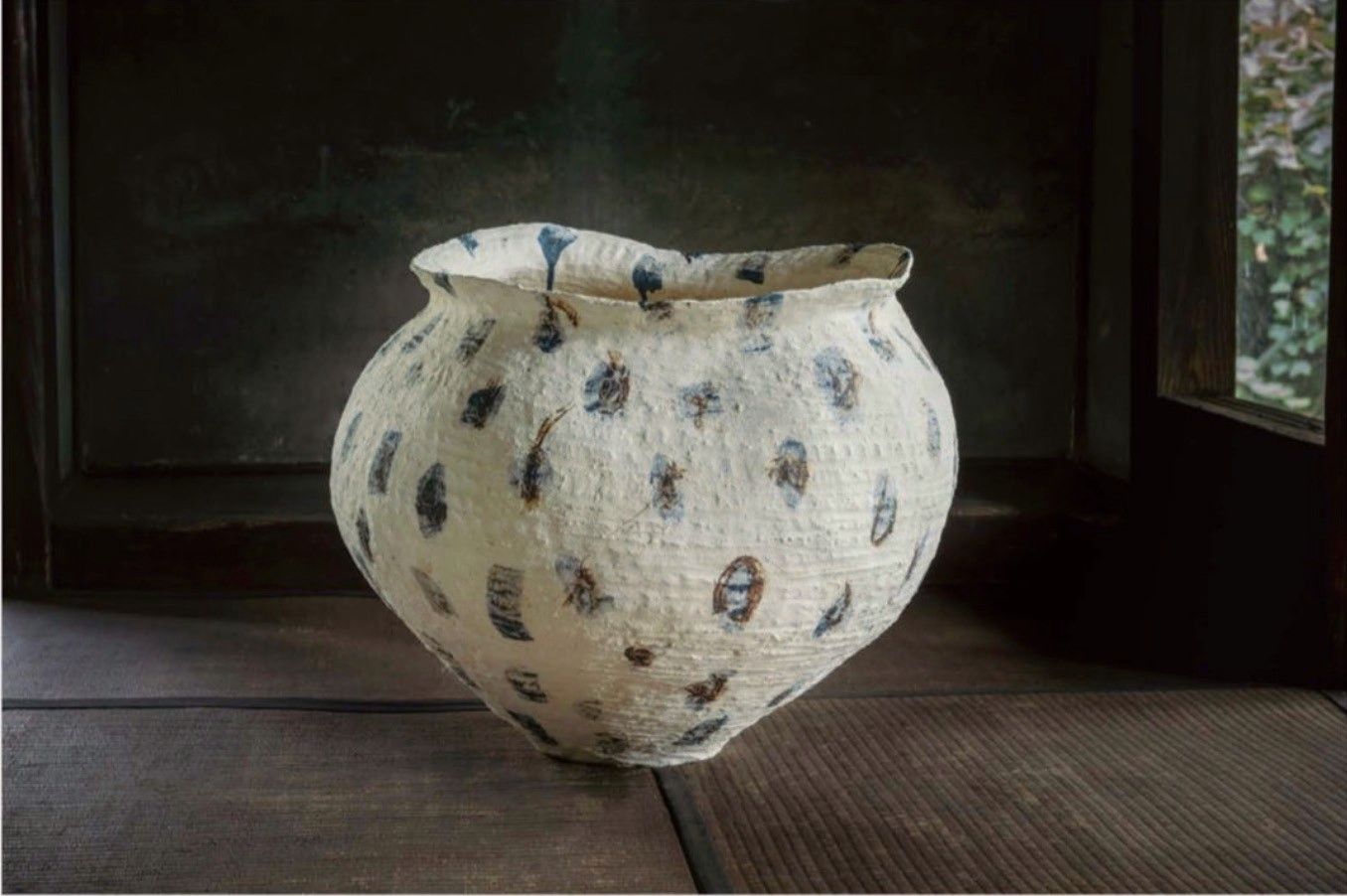 A large white jar with dark speckles on a tatami floor beside a window.