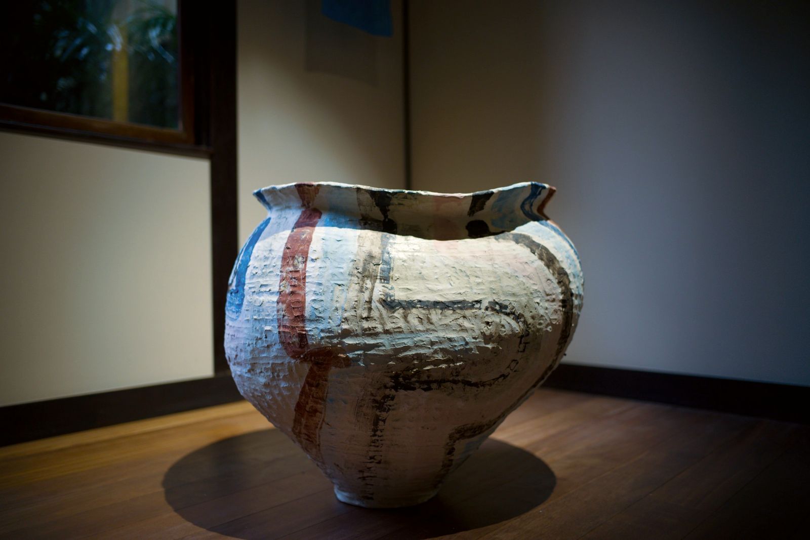 A large hand-built jar with red, blue and black painted lines on a wooden floor.
