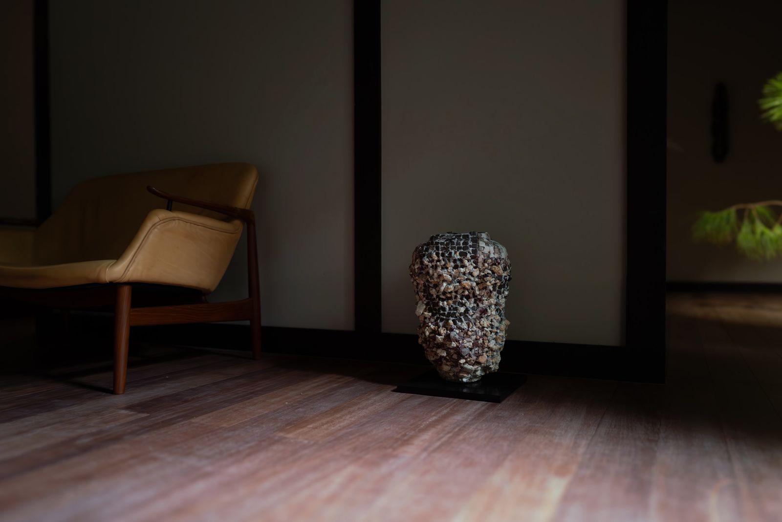 A tall vessel covered in mosaic-like fragments standing on the floor beside a lounge chair.